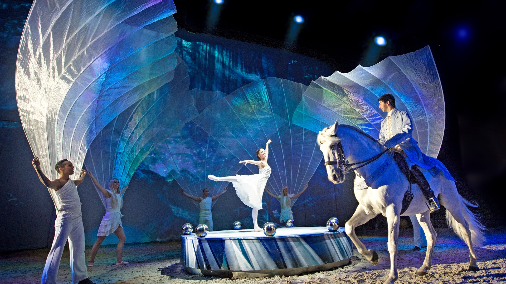 Bühnenszene mit einer Tänzerfigur auf Podest, umgeben von Mitwirkenden mit großen, transparenten Stoffbahnen; daneben reitet eine Person ein weißes Pferd, alles in blauem Bühnenlicht