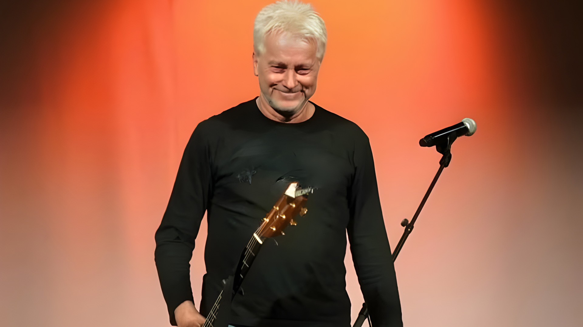 Der Künstler Werner Schmidbauer schwarz gekleidet mit Gitarre und einem Mikrofonständer, der Hintergrund orangerot beleuchtet
