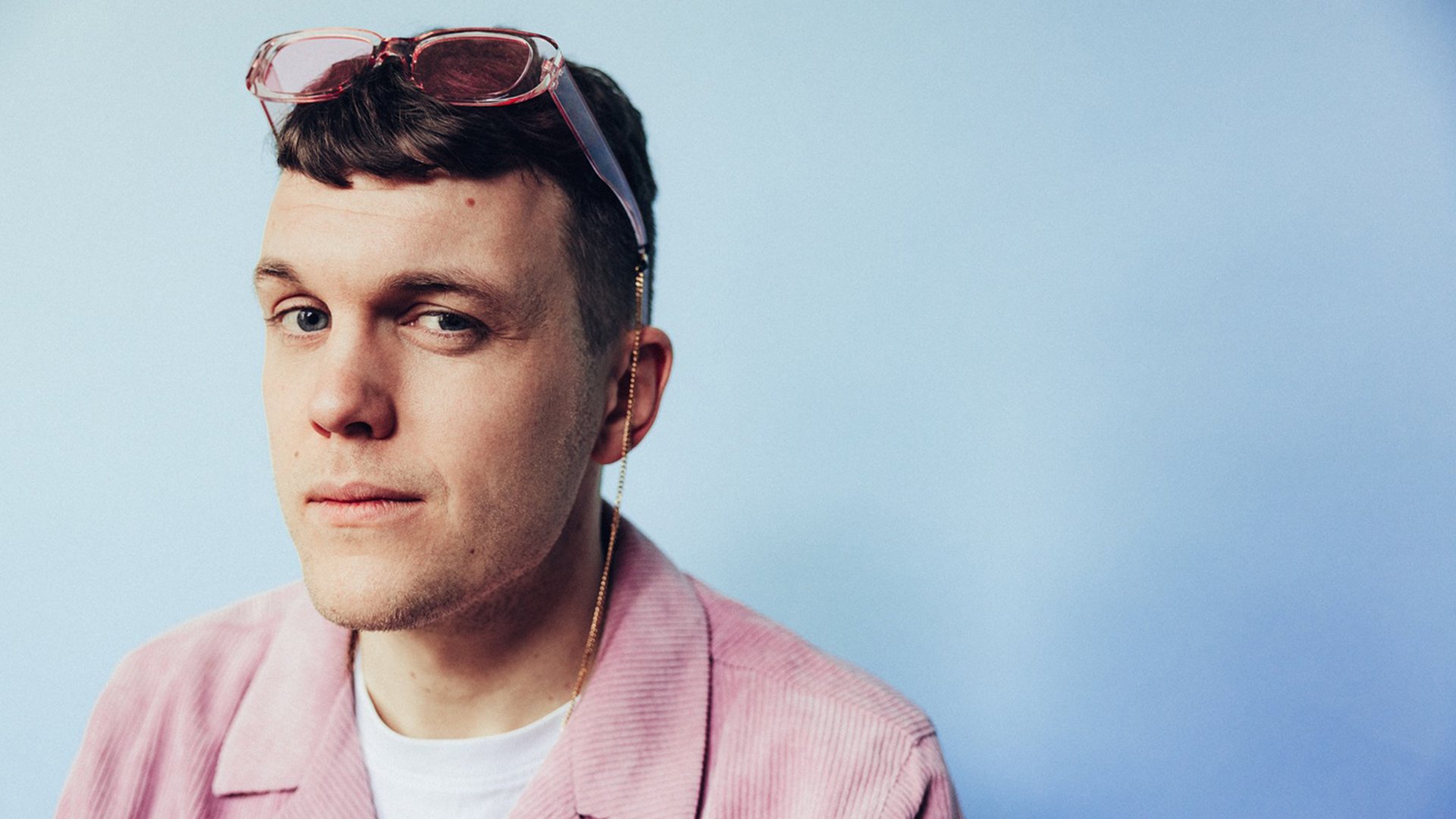 Eine Person in rosa Cordjacke und weißem T‑Shirt trägt eine rosafarbene Brille auf dem Kopf; der Hintergrund ist hellblau und einfarbig.