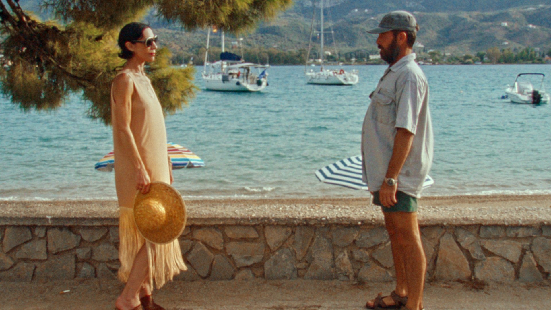 Ein Foto für das Event „KYUKA – BEFORE SUMMER'S END“ bei der Griechischen Filmwoche München: Zwei Personen stehen sich gegenüber und schauen sich an, im Hintergrund das Meer mit einigen Booten und Sonnenschirmen.