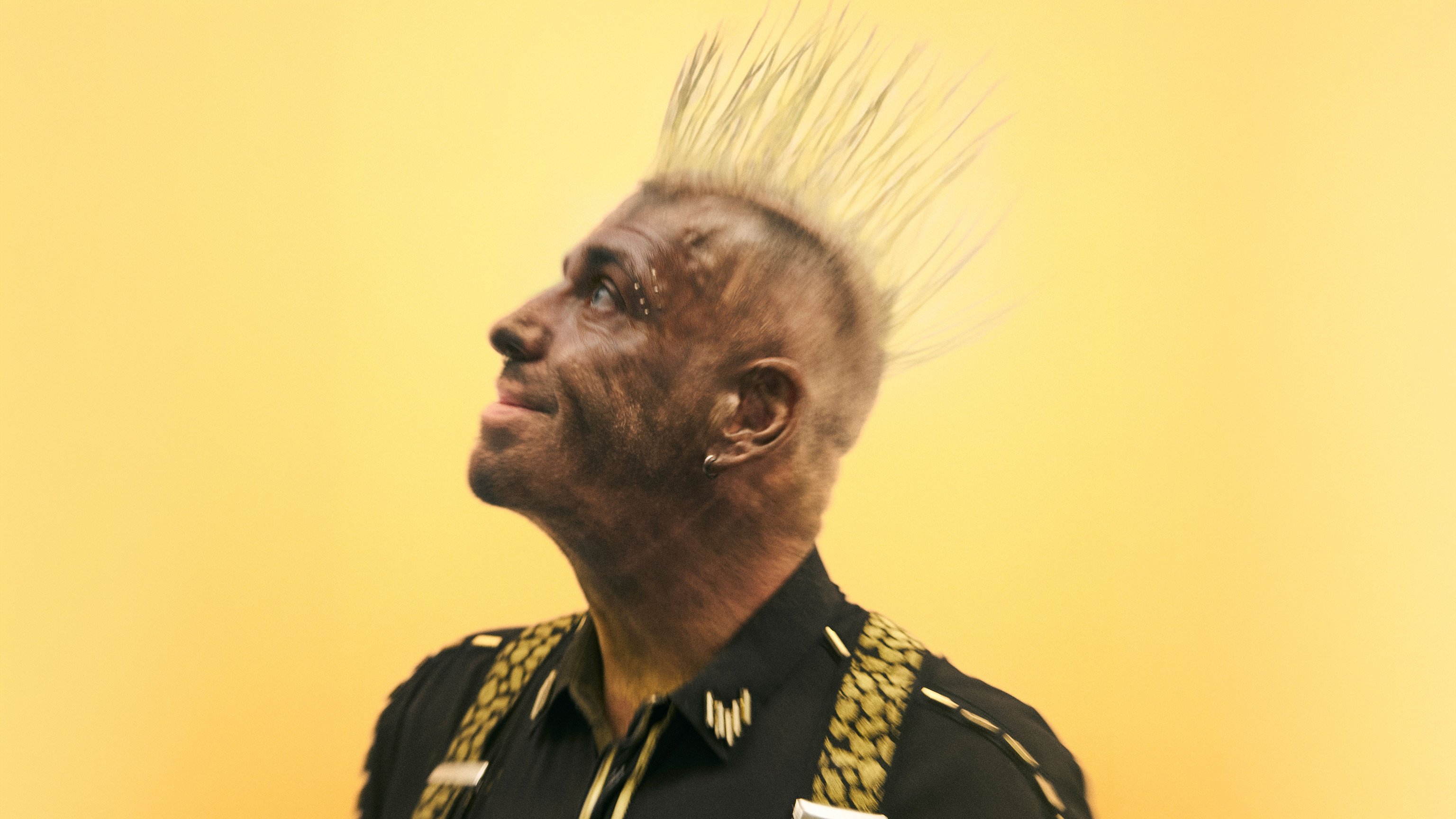 Till Lindemann mit Irokesenfrisur und schwarzem Hemd blickt vor gelbem Hintergrund nach oben. Auffälliges Augen Make up und helle hochstehende Haare prägen die Porträtaufnahme.