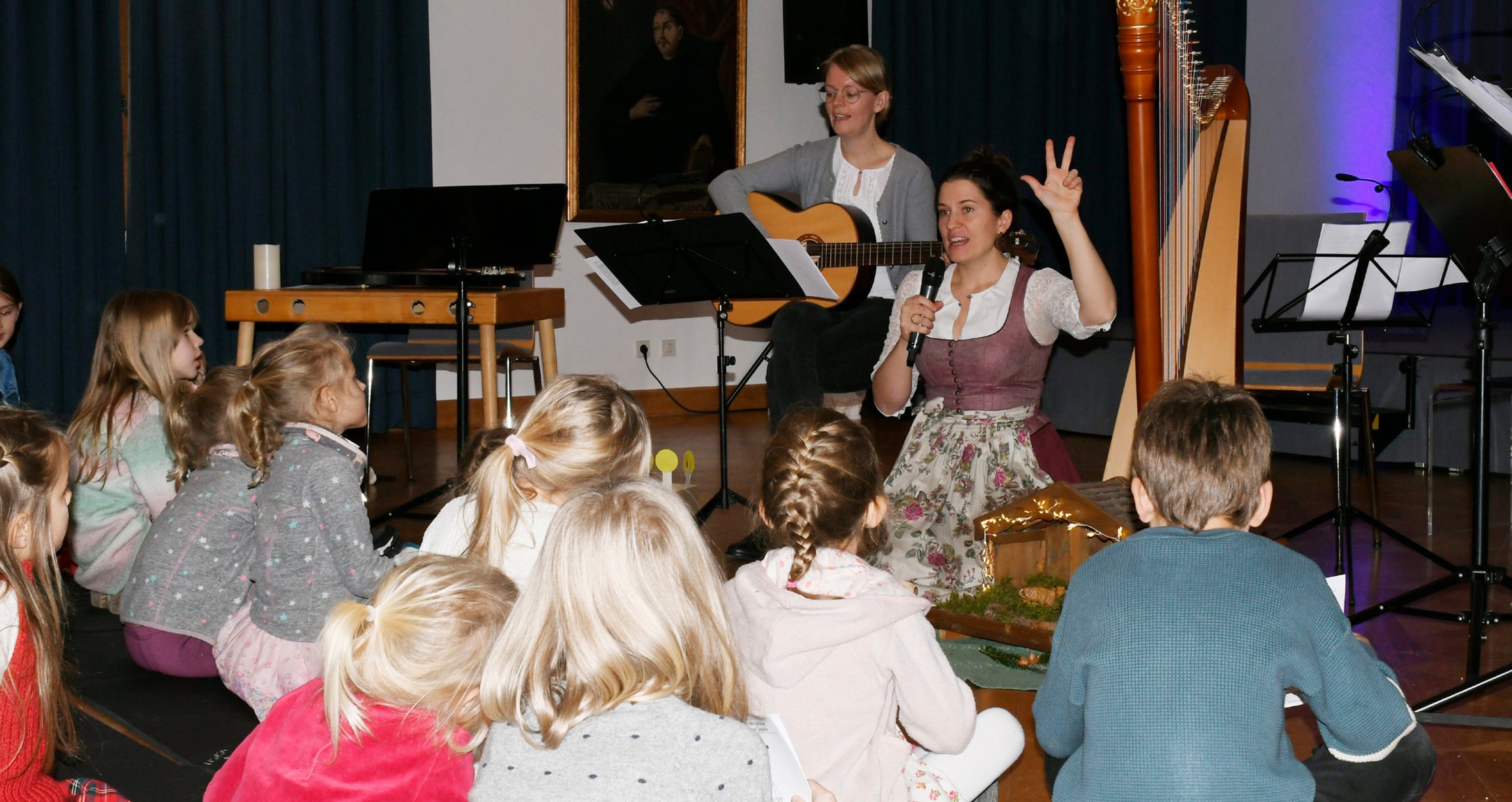 Foto einer Frau mit einer Gitarre, einer weiteren mit einem Mikrofon und einer Gruppe von Kindern.