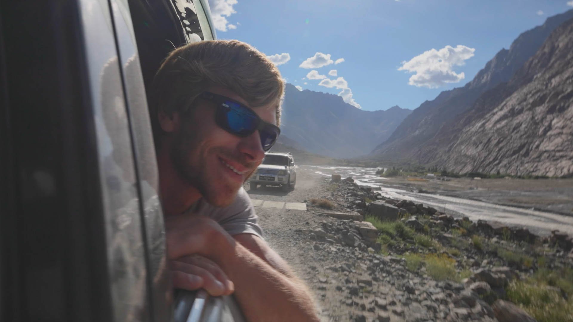 Eine Person lehnt sich aus dem Fenster eines fahrenden Fahrzeugs und stützt die Arme auf dem Türrahmen; die Person trägt eine Sonnenbrille. Im Hintergrund erstreckt sich eine bergige Landschaft mit einem Flussbett oder einer breiten Straße, die sich durch das Tal zieht, unter klarem Himmel im Tageslicht.