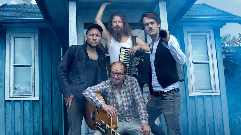 Die vier Künstler des Gankino Circus mit Instrumenten vor einer Holzhütte 