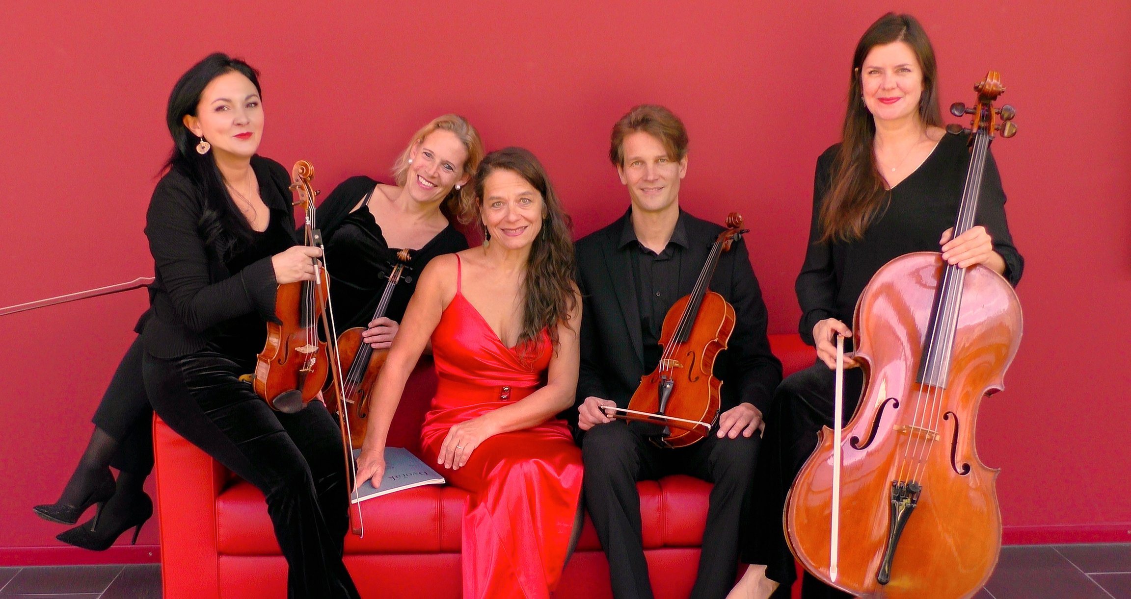 Foto der Gruppe mit Instrumenten, auf einem roten Sofa sitzend, mit einer roten Wand im Hintergrund.