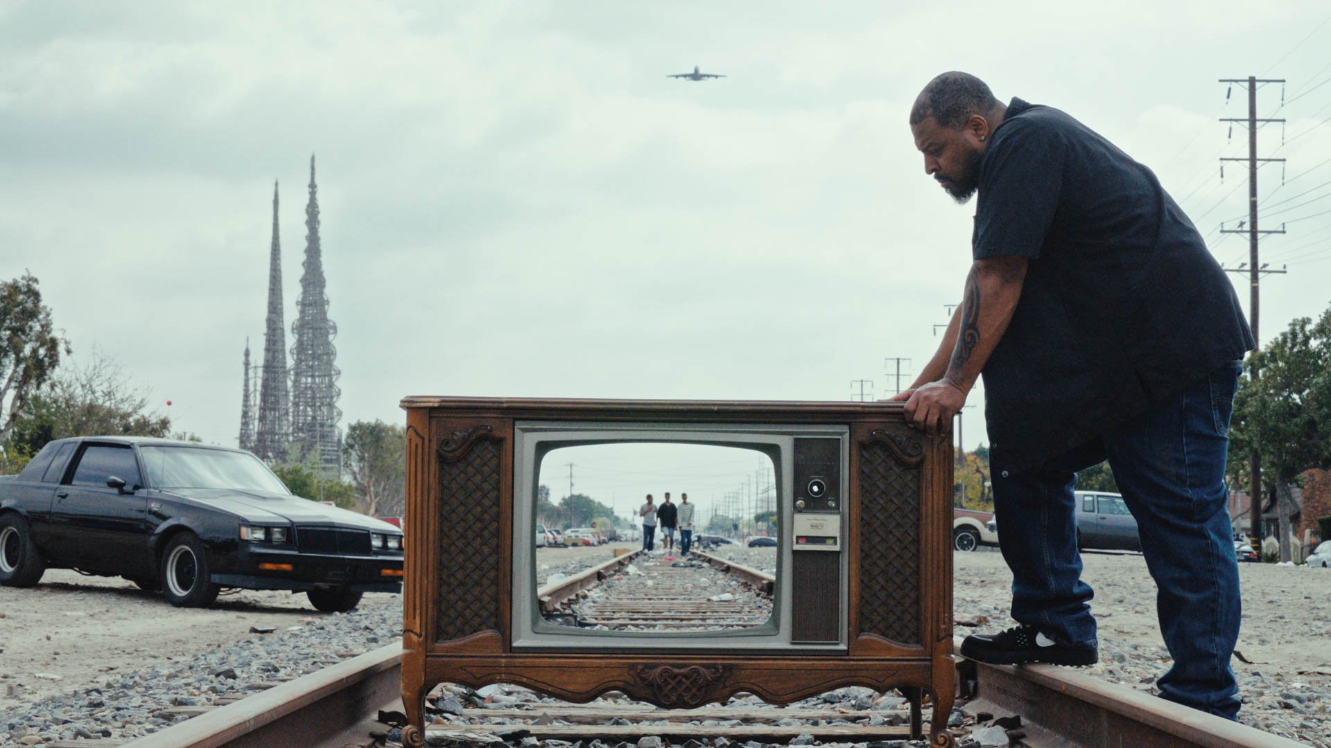 Person schiebt einen alten Fernseher auf Bahngleisen, im Bildschirm ist dieselbe Gleisszene mit zwei Figuren zu sehen, im Hintergrund Autos und Strommasten.