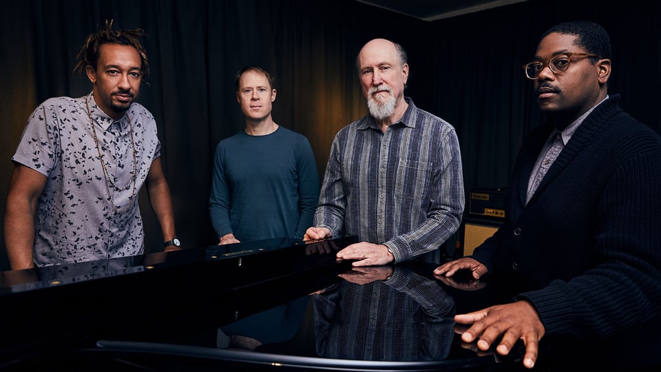 Foto für das Event John Scofield Combo 75 mit der Gruppe, die sich an ein Klavier anlehnt.