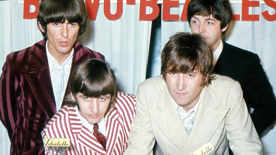 Historisches Pressefoto der Beatles in auffälligen Anzügen vor dem Schriftzug Beatles. Zwei Personen stehen im Hintergrund, während zwei weitere Personen im Vordergrund nach vorne gebeugt in die Kamera blicken.