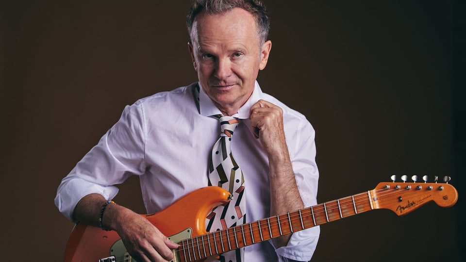 Willy Astor in einem hellblauen Hemd und gemusterter Krawatte mit einer E-Gitarre vor dunklem Hintergrund