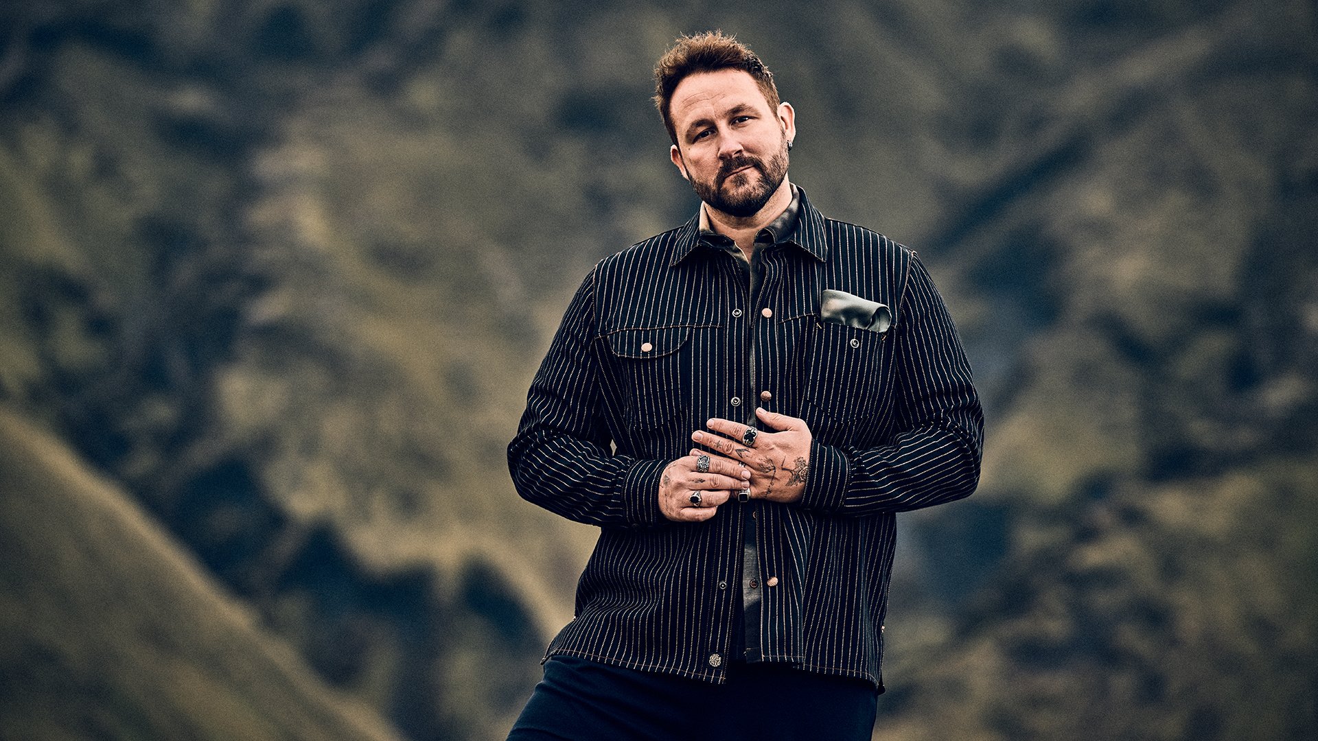 Porträt von Florian Künstler vor unscharfer Berglandschaft, trägt dunkles gestreiftes Hemd mit sichtbaren Ringen und Tattoos.