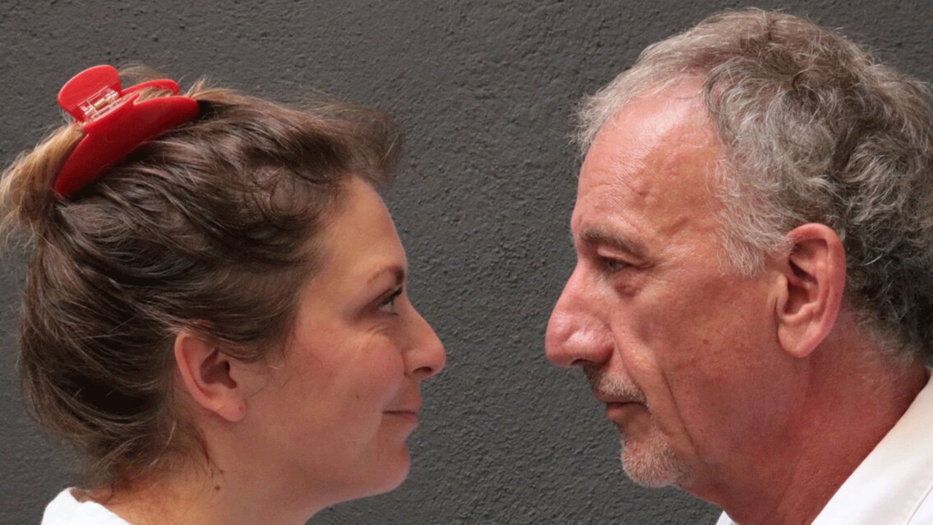 Zwei Personen stehen sich im Profil gegenüber und blicken einander direkt an. Eine Person trägt das Haar hochgesteckt mit einer roten Haarspange, die andere hat kurzes graues Haar und einen grauen Bart.