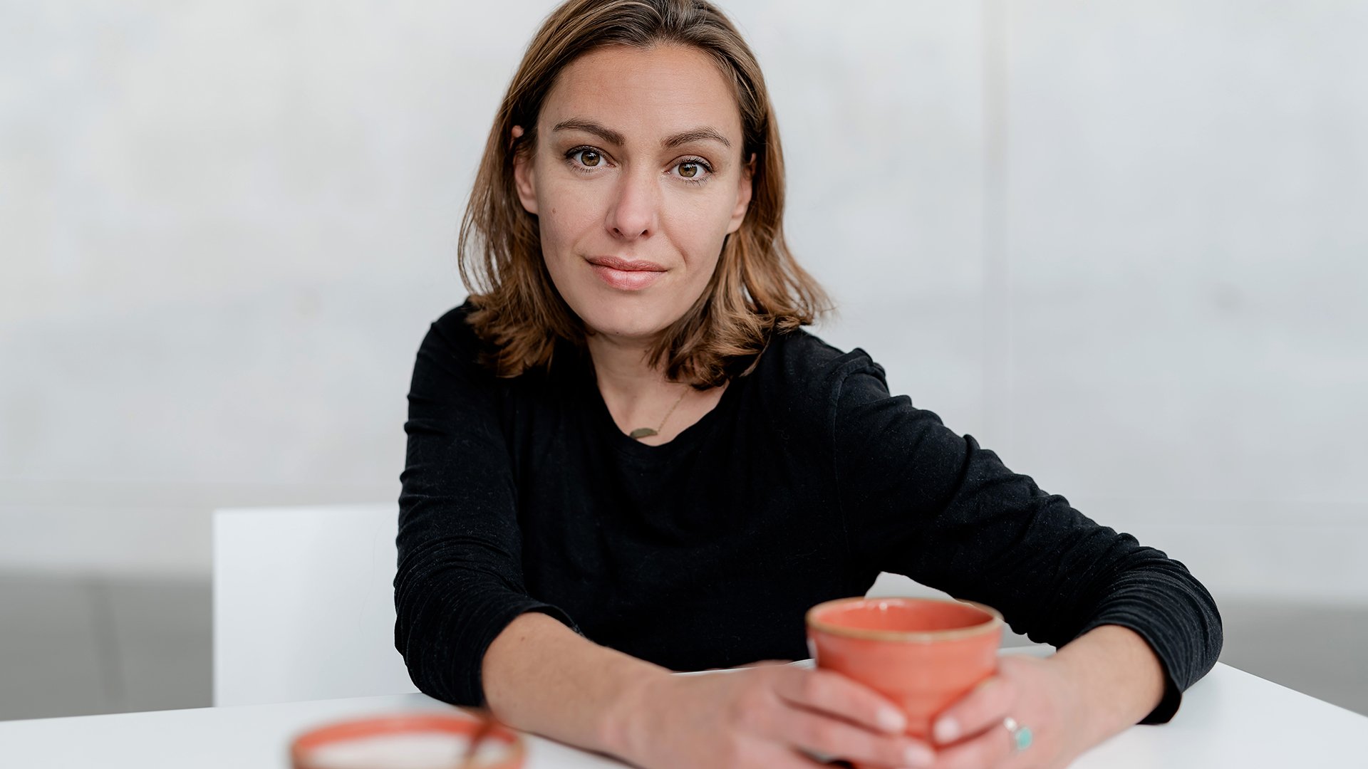 Die Autorin Joana Osman in einem schwarzen Pullover mit einer Tasse in der Hand an einem weißen Tisch sitzend