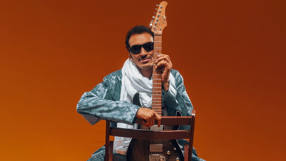 Eventfoto „Bombino“ mit dem Künstler auf einem Stuhl mit Gitarre vor rotem Hintergrund.