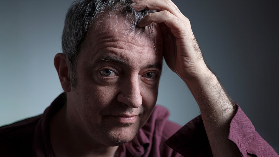 Der Künstler Rainald Grebe in einem dunkelroten Hemd, den Kopf in die Hand gestützt
