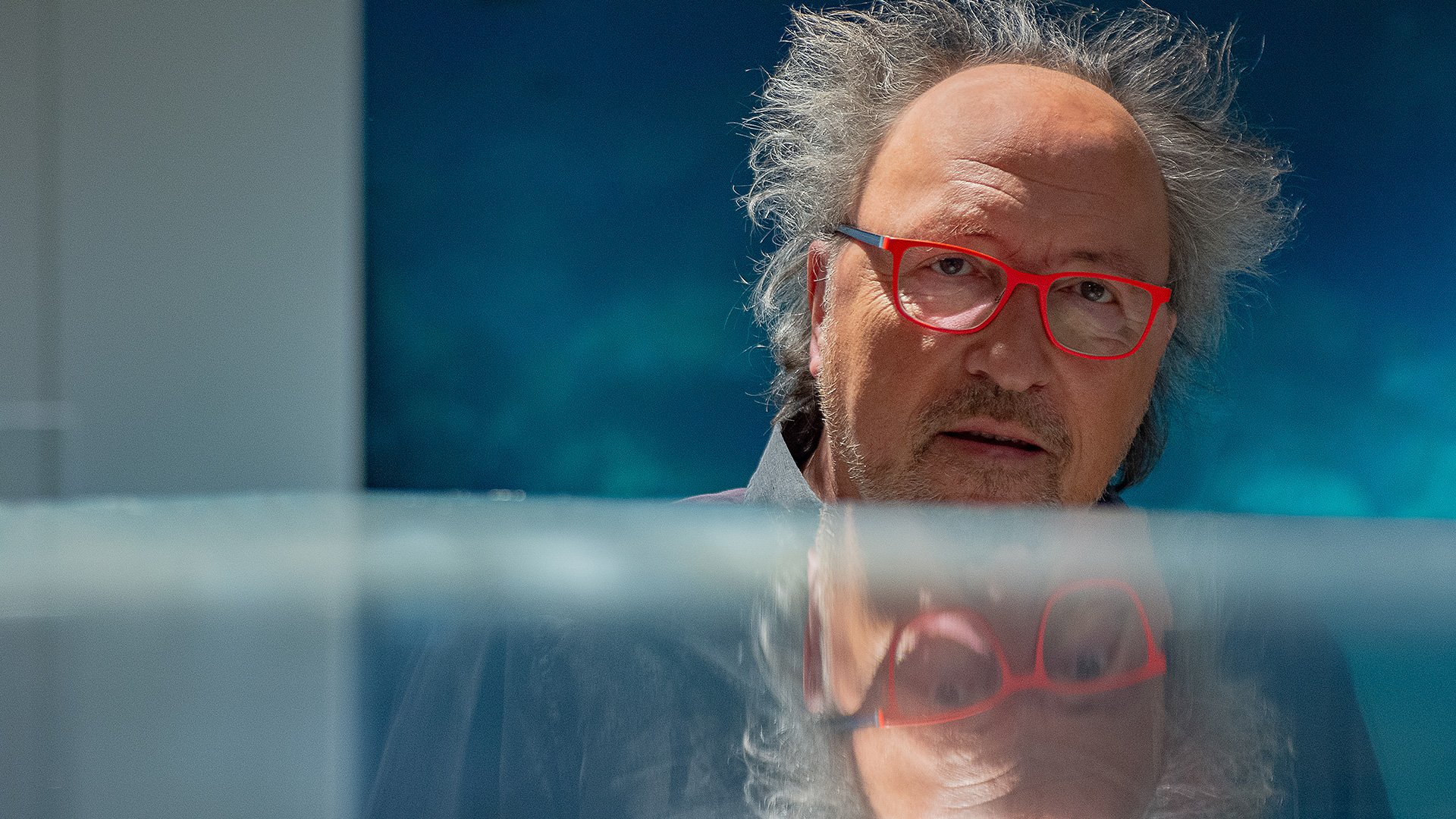 Foto für das Event Urban Priol - Im Fluss mit dem Künstler, der rote Brillen trägt, vor einem blauen Hintergrund.