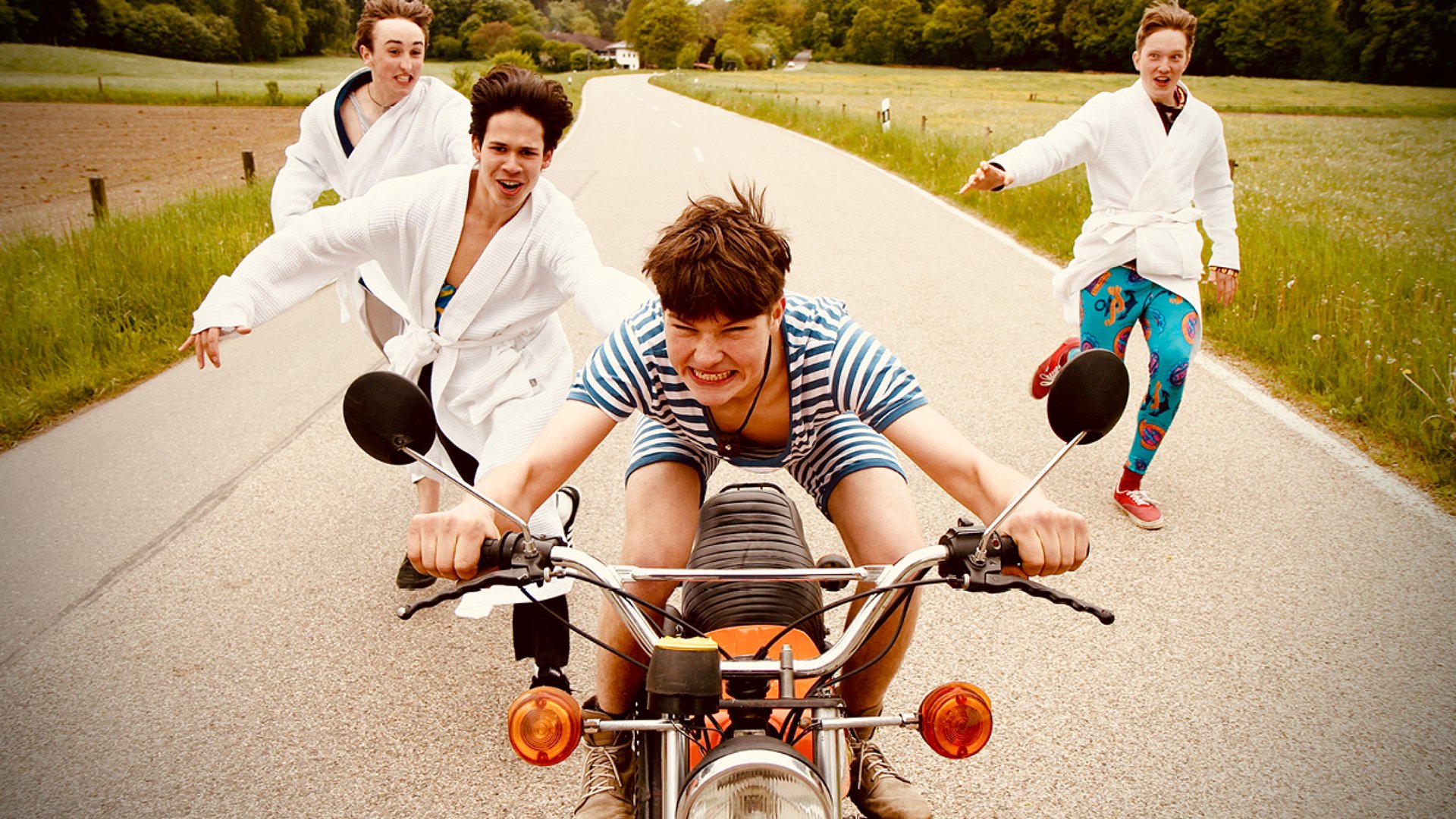 Drei Personen in weißem Bademantel laufen hinter einem Motorrad mit einer vierten Person in gestreifter Kleidung her