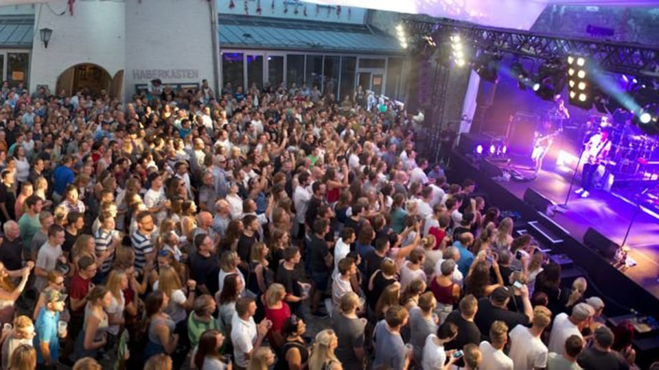 Große Menschenmenge bei Open-Air-Konzert vor Bühne mit Lichtshow, dichtes Publikum und Festivalatmosphäre in urbanem Innenhof
