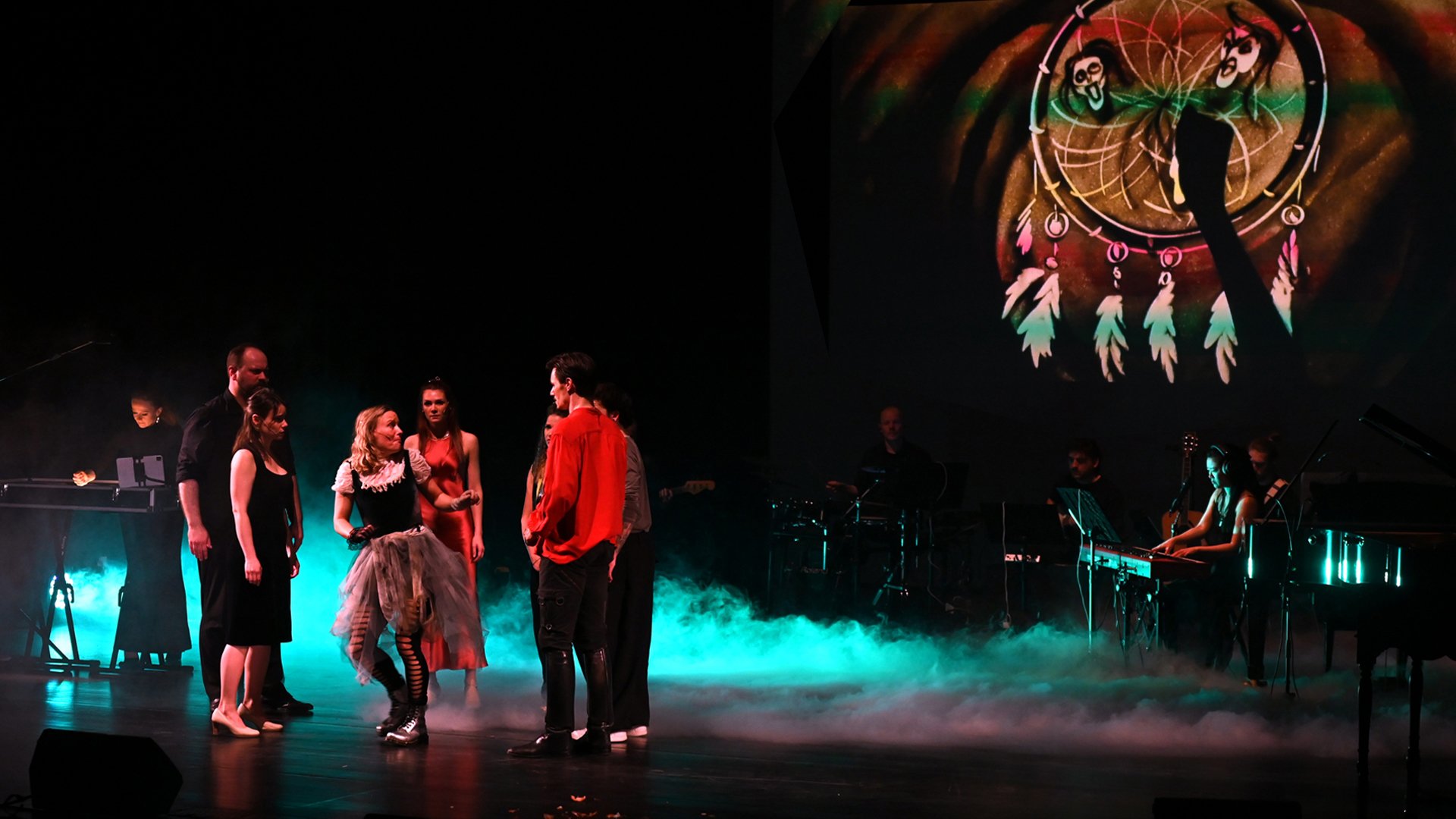 Bühnenaufnahme eines Musical-Ensembles mit mehreren Darstellern im Vordergrund und Live-Band im Hintergrund; farbiges Licht, Nebeleffekte und eine projizierte Traumfänger-Grafik schaffen eine atmosphärische Szene während der Aufführung.