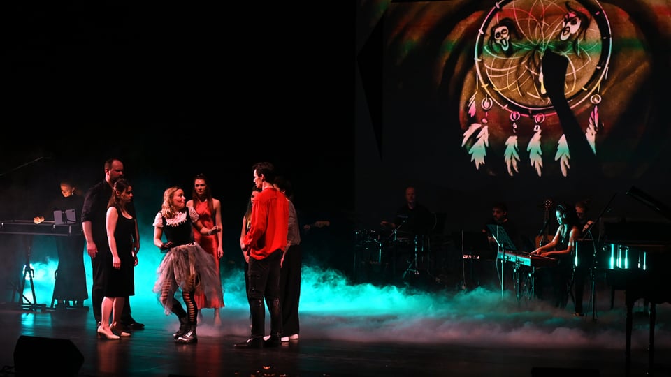 Bühnenaufnahme eines Musical-Ensembles mit mehreren Darstellern im Vordergrund und Live-Band im Hintergrund; farbiges Licht, Nebeleffekte und eine projizierte Traumfänger-Grafik schaffen eine atmosphärische Szene während der Aufführung.