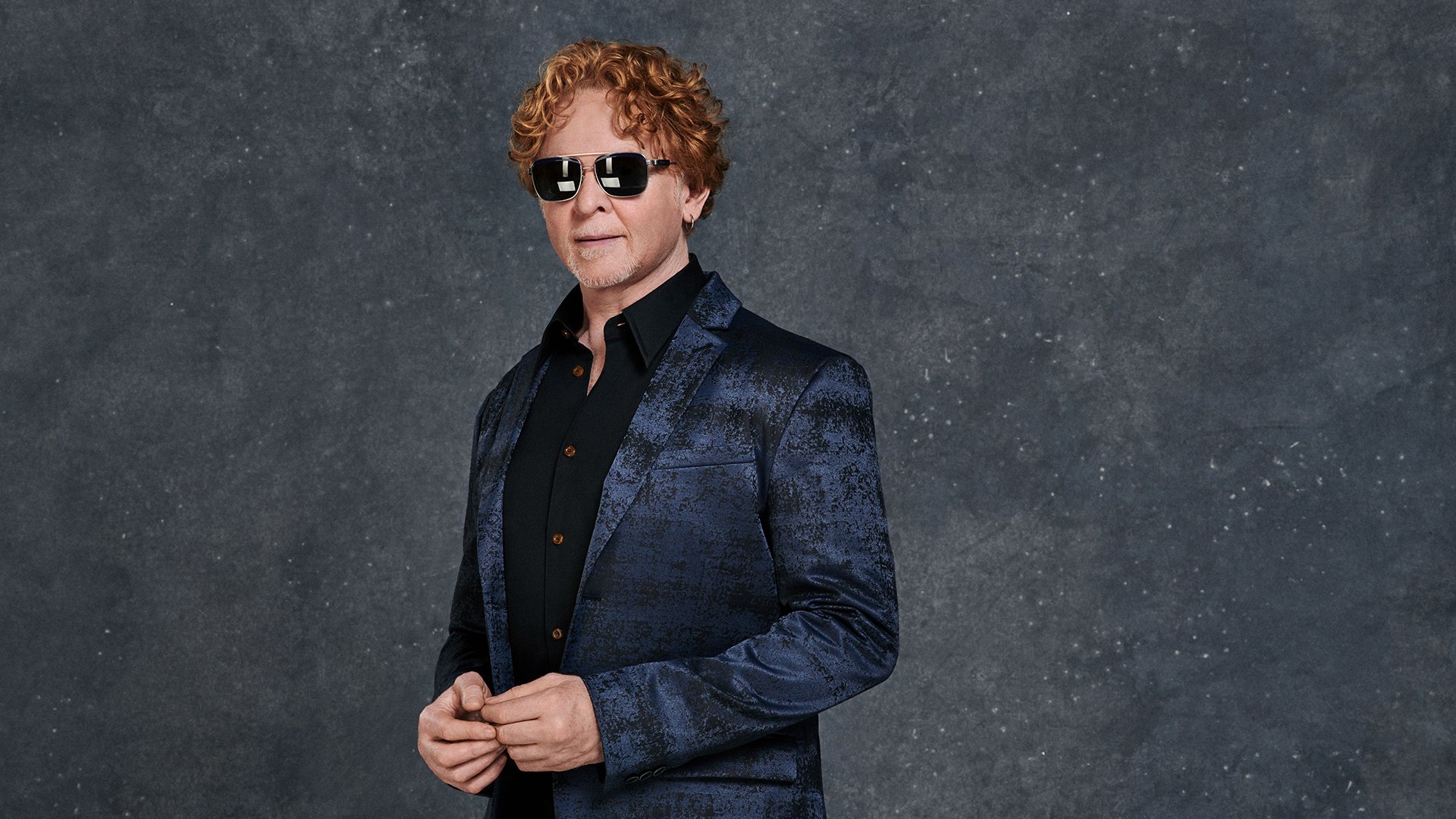 Porträtfoto von Mick Hucknall in dunkelblauem glänzendem Sakko und schwarzem Hemd vor grauem Hintergrund, mit Sonnenbrille und lockigem rotem Haar