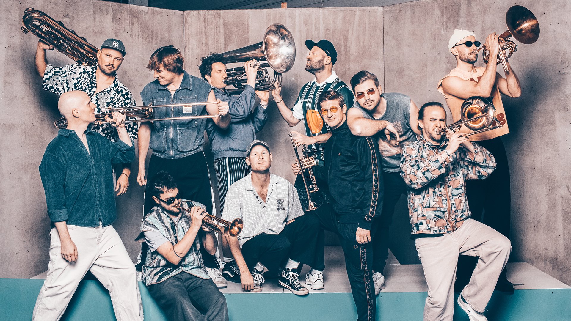 Bandfoto von Querbeat: Mehrere Personen mit Blechblasinstrumenten (u. a. Trompete, Posaune, Tuba) gruppieren sich auf einer erhöhten Plattform vor betonfarbener Wand. Unterschiedliche Posen mit Instrumenten im Vorder‑ und Hintergrund, legere Bühnenkleidung. Kühles, gleichmäßiges Licht, dynamische Gesamtkomposition; Promomotiv zur Live‑Show von Querbeat beim Tollwood Sommerfestival in München.