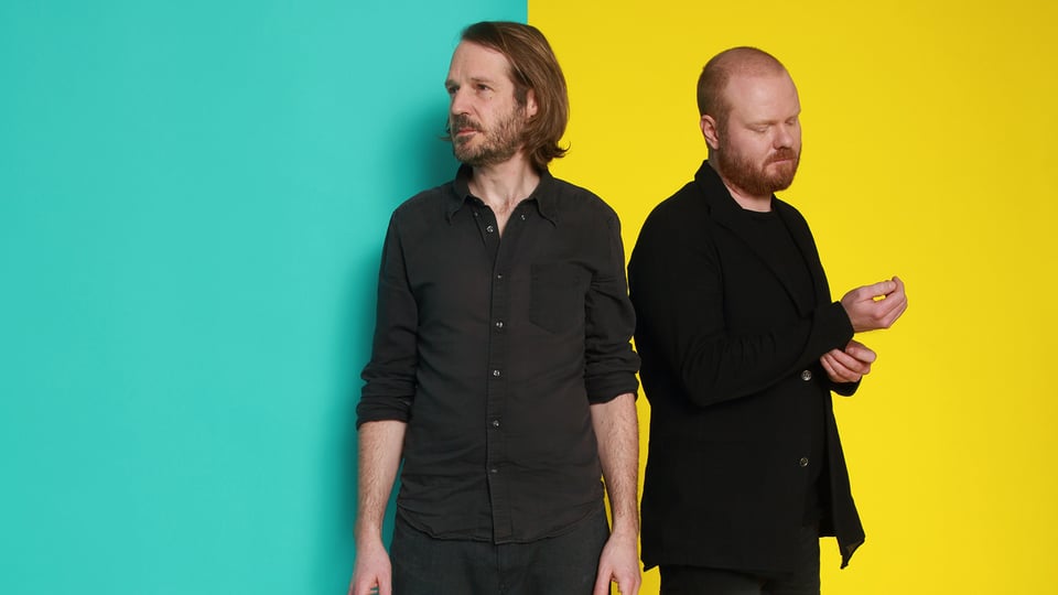 Foto für die Veranstaltung Gisbert zu Knyphausen und Kai Schumacher, mit beiden Künstlern vor einem Hintergrund, der zur Hälfte gelb und zur Hälfte türkis ist.