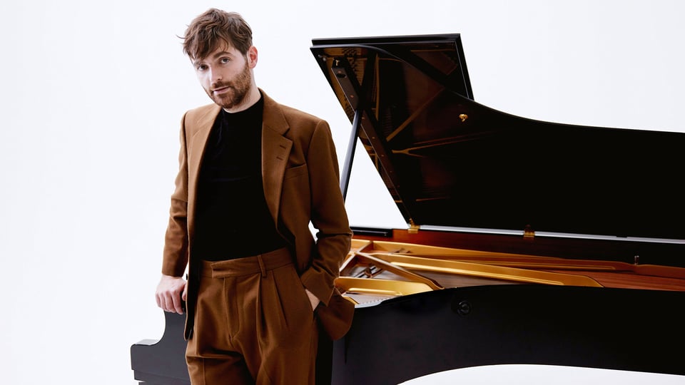 Foto von Florian Christl mit dem Künstler und einem Piano im Hintergrund.