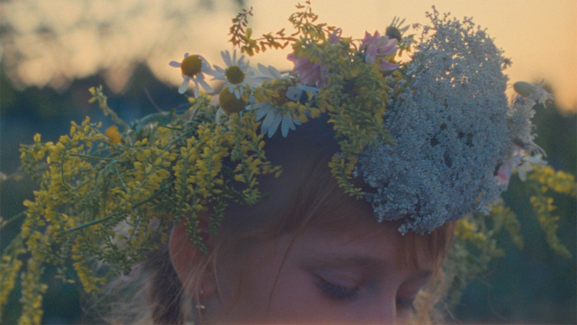 Nahaufnahme einer Person im Freien, deren Kopf mit einem Kranz aus verschiedenen Blumen und Pflanzen bedeckt ist; die Person blickt nach unten, während das warme, weiche Licht des Sonnenuntergangs die Szene umgibt und der Hintergrund unscharf bleibt.