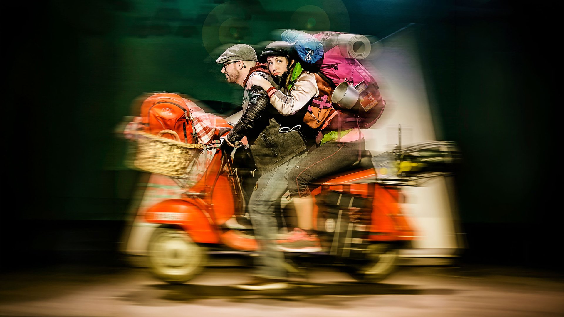 Zwei Personen fahren auf einem roten Roller mit Gepäck durch eine Szene, mit Bewegungsunschärfe und Bühnenlicht im Hintergrund.