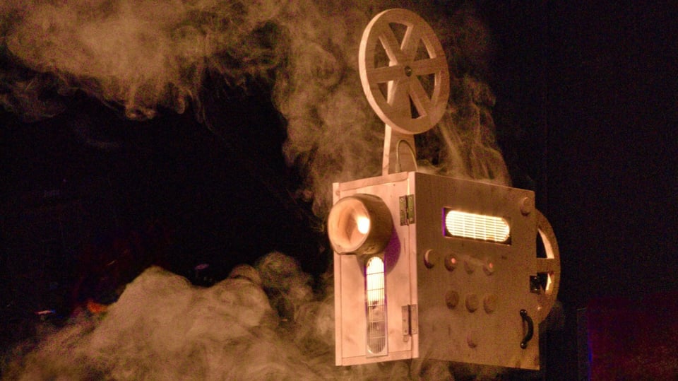 Bühnenbild mit einer beleuchteten Filmprojektor-Requisite, aus der Rauch aufsteigt, vor dunklem Hintergrund.