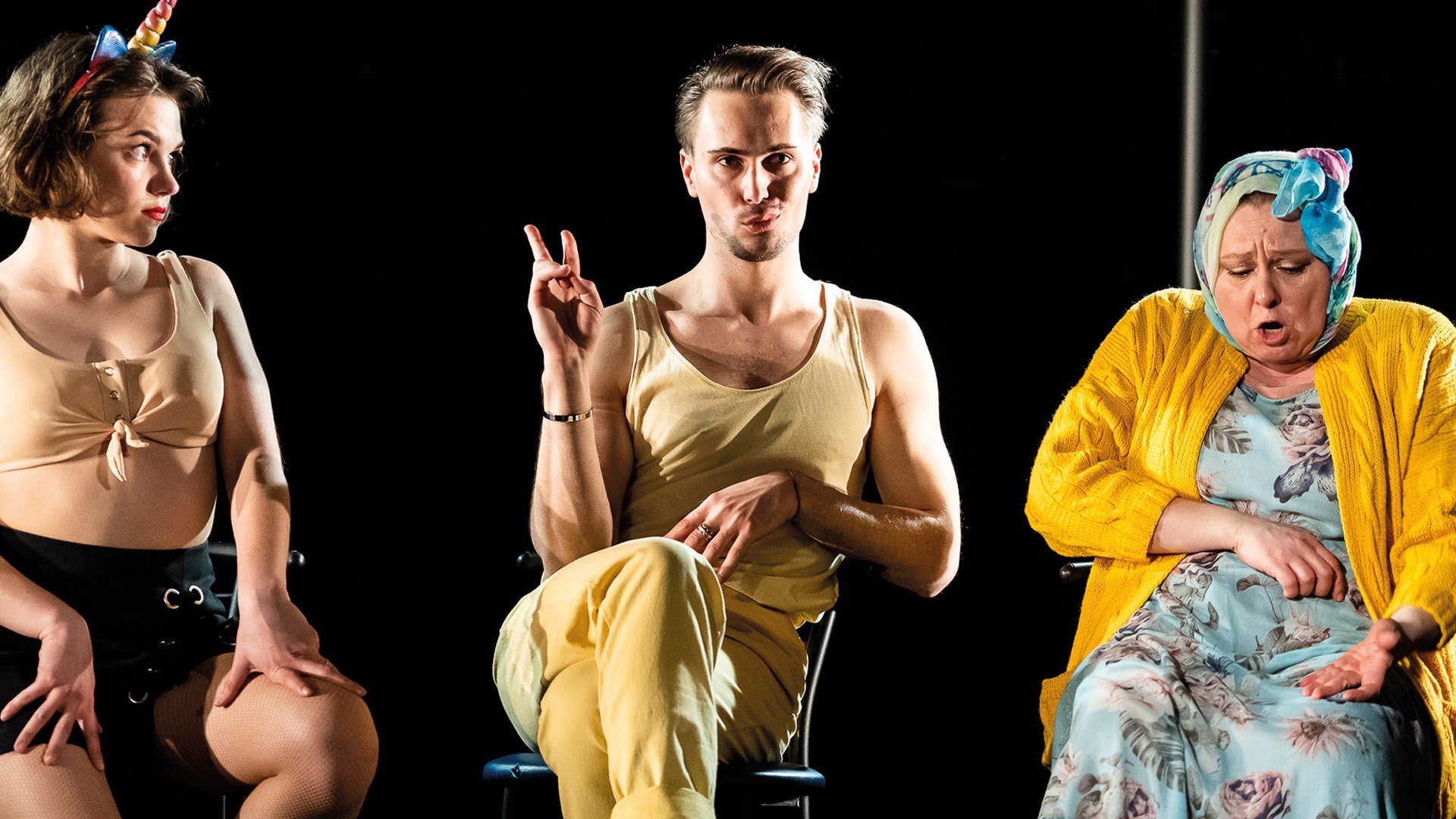 Drei Personen in unterschiedlichen Bühnenkostümen sitzen nebeneinander auf einer Theaterbühne vor schwarzem Hintergrund. Die Person in der Mitte trägt ein helles Outfit und sitzt mit übergeschlagenen Beinen auf einem Stuhl, während die beiden anderen Personen farbenfrohe Kostüme und Kopfbedeckungen tragen. Die Szene wird von hellem Bühnenlicht beleuchtet.