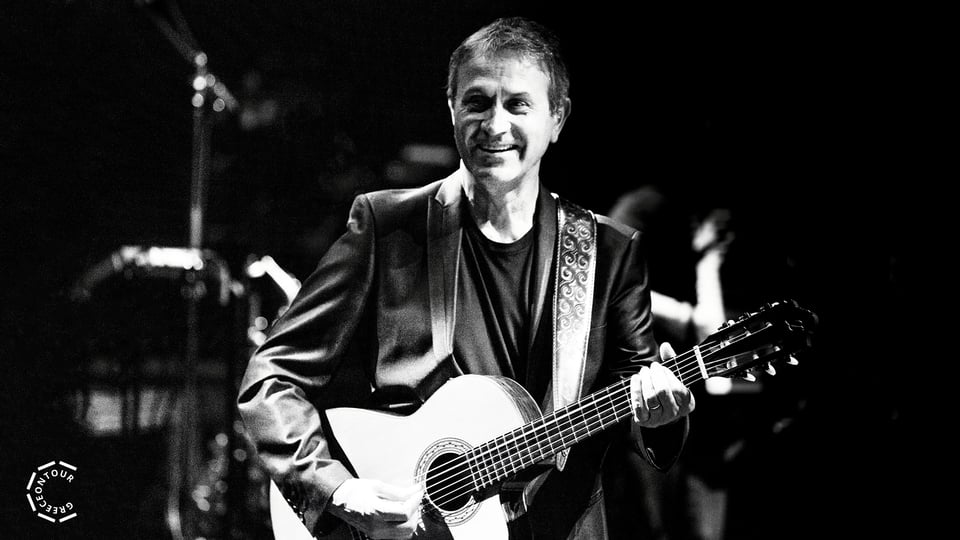 Person spielt eine akustische Gitarre auf einer Bühne in Schwarzweißaufnahme; im Hintergrund sind Schlagzeug und Bühnenlichter leicht unscharf sichtbar.