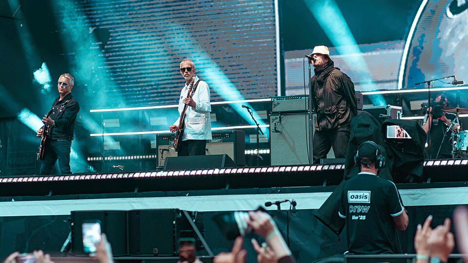 Liveauftritt der Band Oasis auf einer Festivalbühne. Mehrere Musiker stehen mit Gitarren und Mikrofonen unter blauen Lichtstrahlen, während im Vordergrund Publikum und ein Crewmitglied vor der Bühne zu sehen sind.