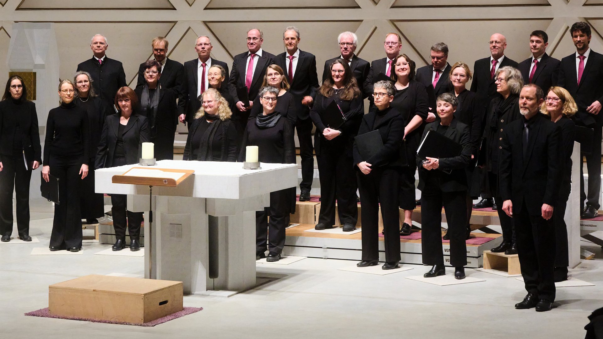 Gruppenfoto des Chors Vox Nova mit zahlreichen Chormitgliedern in schwarzer Konzertkleidung auf Podesten in einer modernen Kirche, vor einem weißen Altar mit Kerzen und geometrisch gestalteter Rückwand.