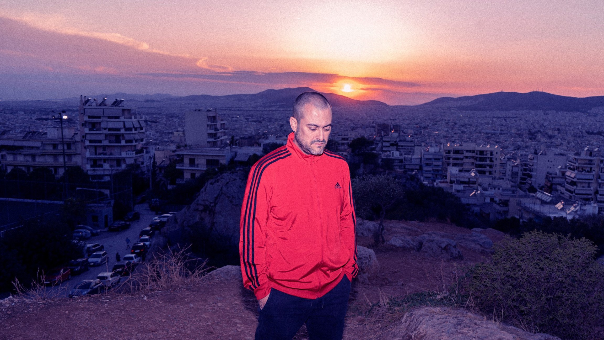 Person in roter Trainingsjacke steht auf einem Hügel vor einer dicht bebauten Stadtlandschaft. Die Sonne geht hinter den Bergen am Horizont unter und taucht Himmel und Stadt in violette und orangefarbene Farbtöne. Felsen, trockene Vegetation und parkende Autos im Vordergrund.
