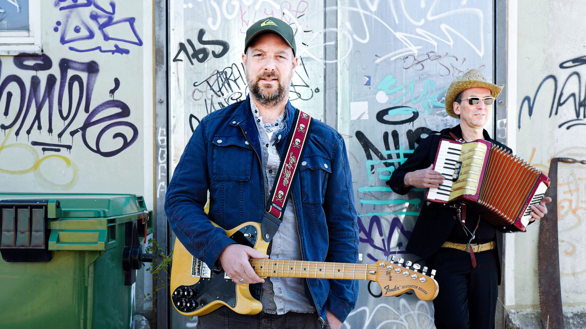 Zwei Musiker vor einer mit Graffiti besprühten Wand. Im Vordergrund steht eine Person mit E-Gitarre und Kappe, im Hintergrund spielt eine Person mit Hut und Sonnenbrille Akkordeon neben einem grünen Müllcontainer.