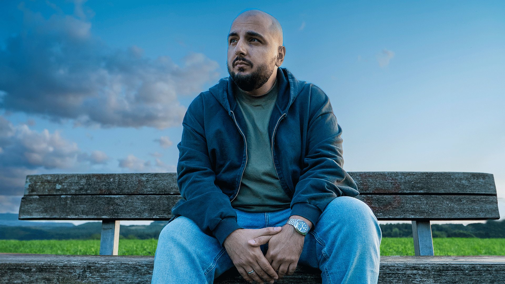 Künstler Sezer Oglu sitzt auf einer Bank im Freien und blickt in die Ferne vor einer grünen Landschaft und bewölktem Himmel