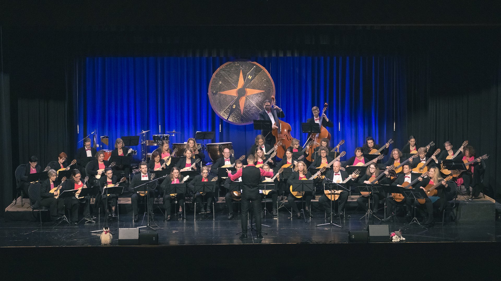 Die MusikerInnen des Vivaldi Orchesters Karlsfeld auf der Bühne in schwarzer Kleidung mit Instrumenten vor einem blauen Vorhang mit einem runden großen Emblem