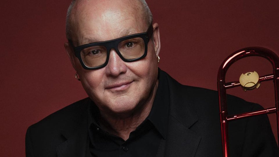 Porträt eines Mannes mit Brille vor rotem Hintergrund, rechts neben ihm eine Posaune; der Musiker blickt frontal in die Kamera.