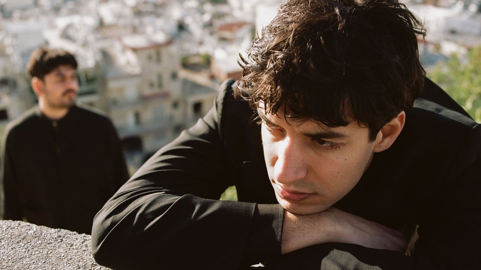 Eventfoto „Apsilon“ mit dem Künstler an einer kleinen Steinmauer, im Hintergrund ein weiterer Mann mit Stadtkulisse.