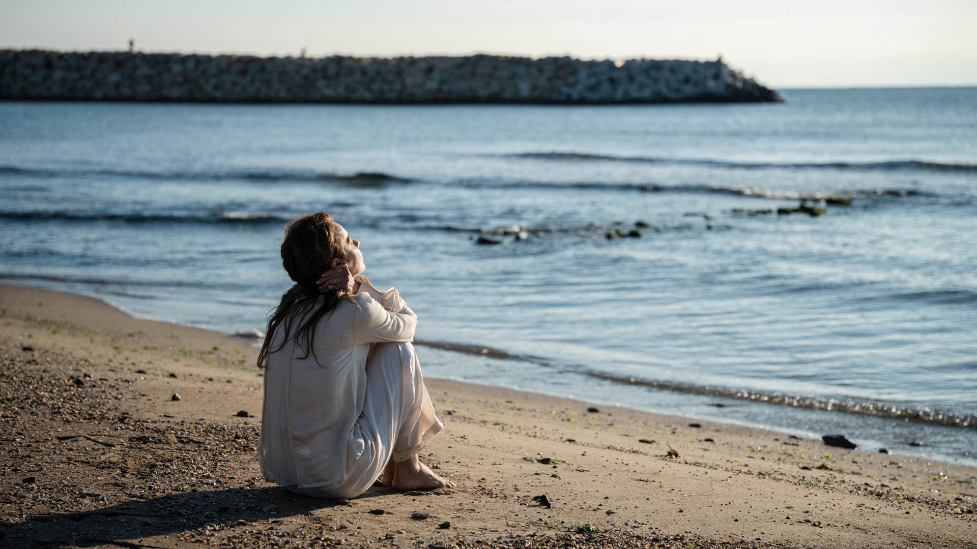 Person sitzt allein am Strand und blickt auf das Meer, im Hintergrund Wellen und eine steinerne Mole.