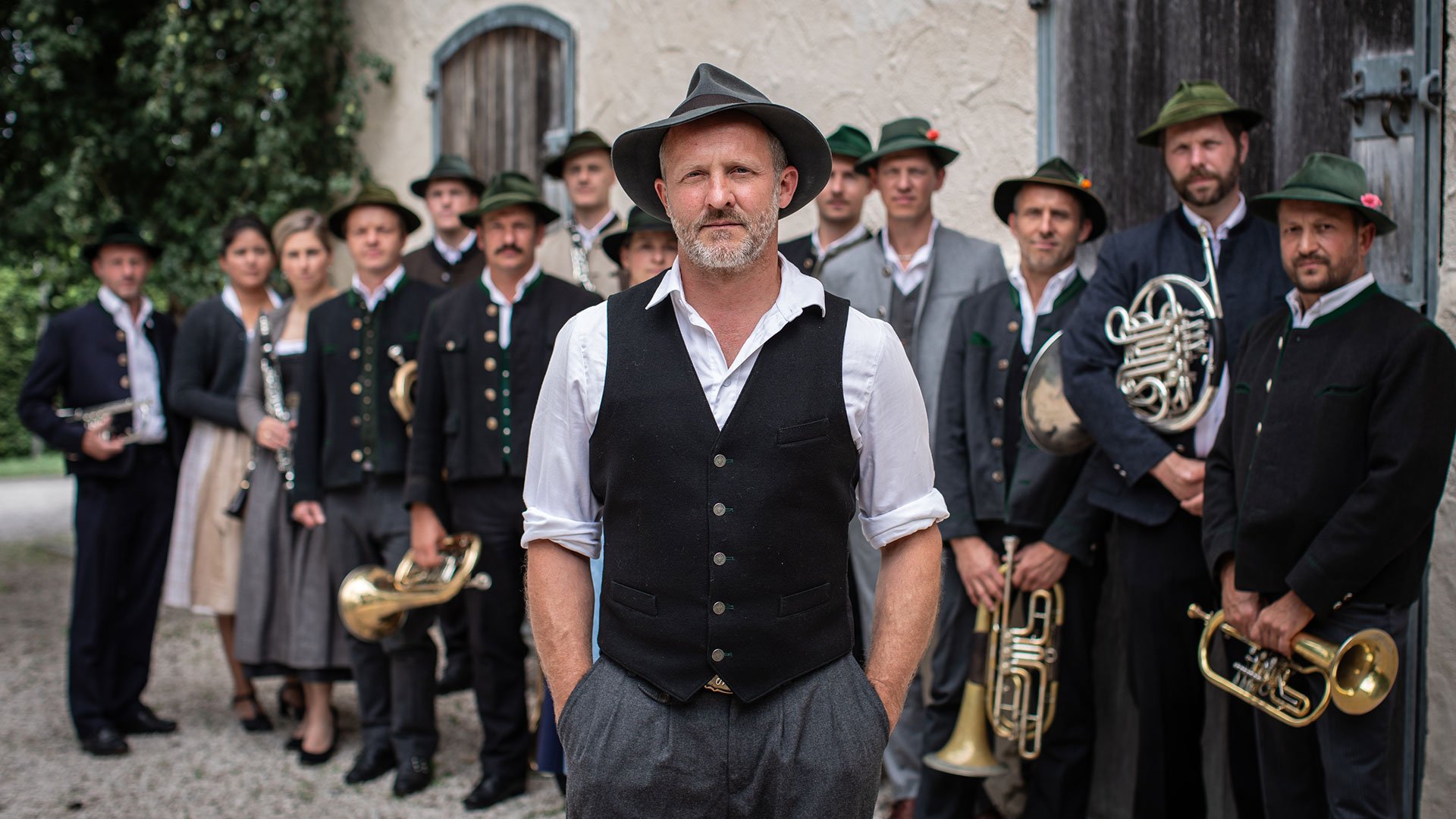 Maximilian Brückner & Die Riederinger: Gruppe von Musikerinnen und Musikern in traditioneller Tracht mit Blasinstrumenten, stehend vor einem Gebäude im Freien; im Vordergrund ein Mann mit Hut und Weste.