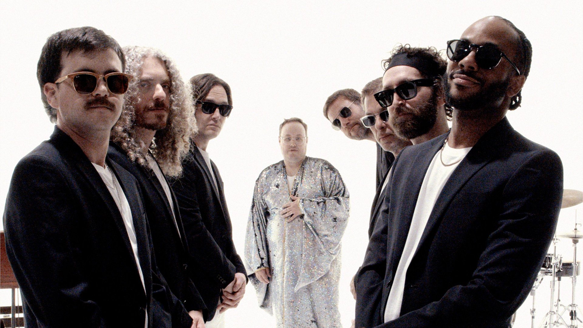 Gruppenfoto von St. Paul & the Broken Bones in einem hellen Studio; mehrere Bandmitglieder stehen seitlich in dunkler Kleidung im Vordergrund, während eine Person in einem glitzernden, weiten Outfit mittig im Hintergrund steht; rechts sind Teile eines Schlagzeugs sichtbar