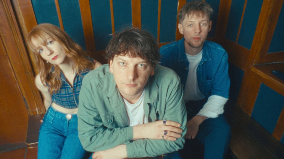 Naked Cameo Bandfoto: Drei Musiker sitzen eng beieinander in einem holzverkleideten Raum, schauen direkt in die Kamera, lässiger Indie-Rock-Look.