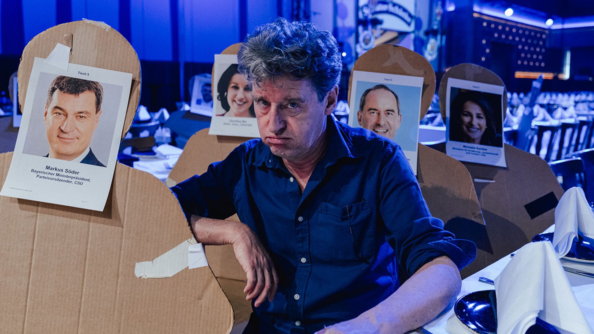 Der Darsteller sitzt zwischen Pappaufstellern mit Bildern von Politikern