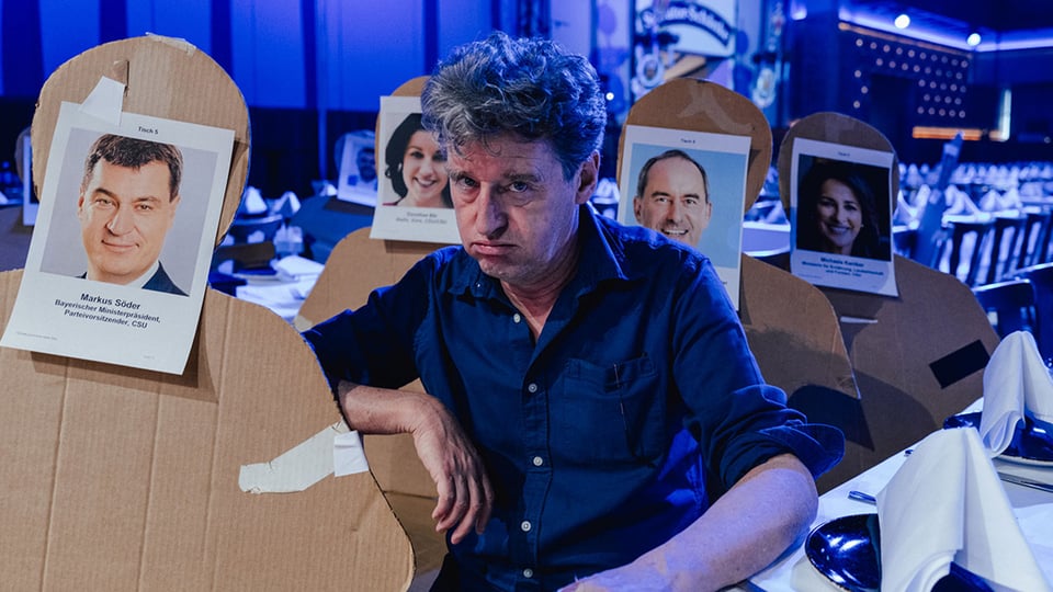 Der Darsteller sitzt zwischen Pappaufstellern mit Bildern von Politikern