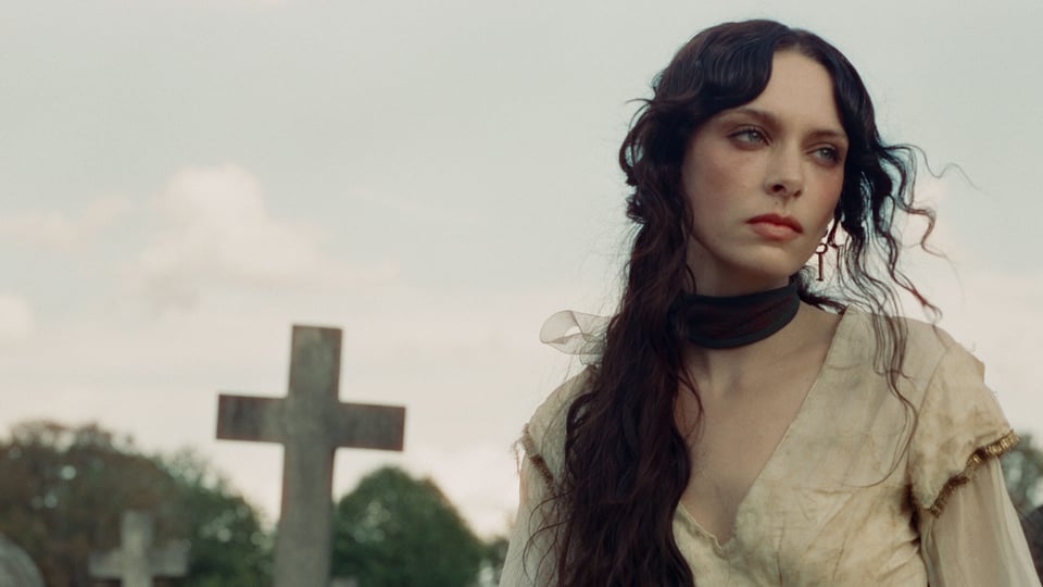 Porträt von Holly Humberstone im Freien, mit langem dunklem Haar und hellem Kleid, im Hintergrund ein unscharfes Kreuz und Bäume.