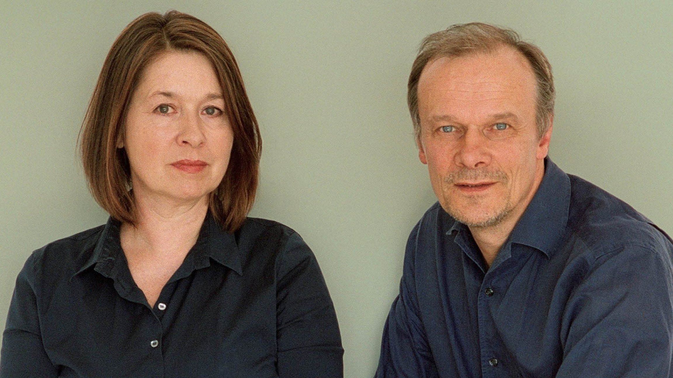 Edgar Selge und Franziska Walser in dunkler Kleidung sitzen nebeneinander vor neutralem Hintergrund und blicken in die Kamera.