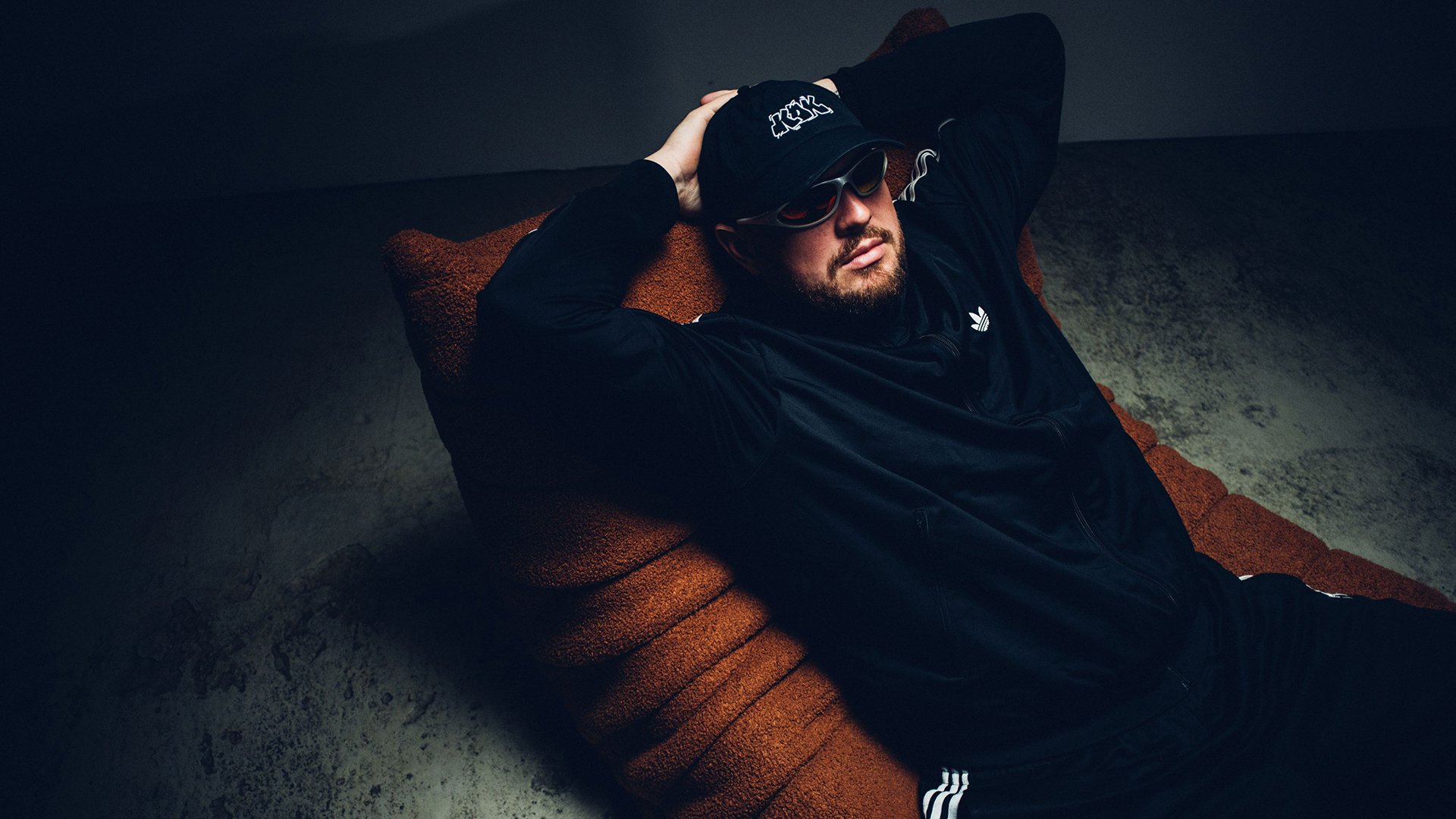 Eine Person in schwarzem Trainingsanzug, mit Sonnenbrille und Kappe, die auf einem braunen Sessel lehnt, vor dunklem Hintergrund