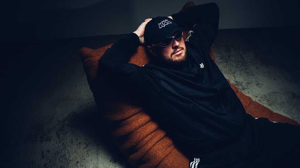 Eine Person in schwarzem Trainingsanzug, mit Sonnenbrille und Kappe, die auf einem braunen Sessel lehnt, vor dunklem Hintergrund